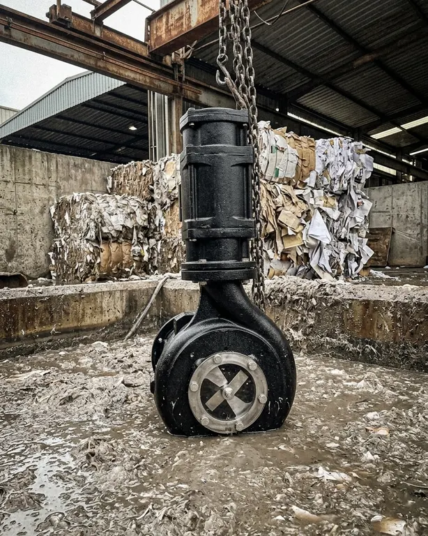 A black chopper pump on its chain in a pit of fibrous pulp water, bales of waste paper stacked behind